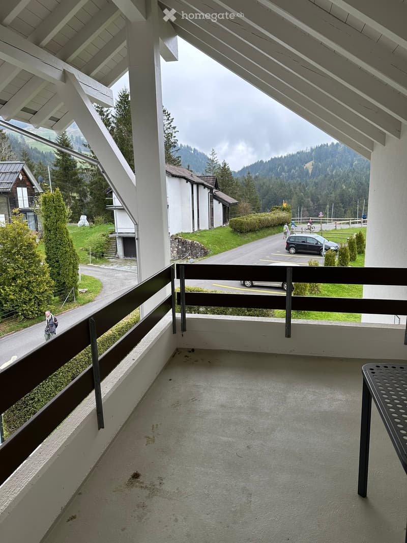 Balcony with a concrete floor, metal railing, and a view of a road, parked cars, and surrounding greenery.