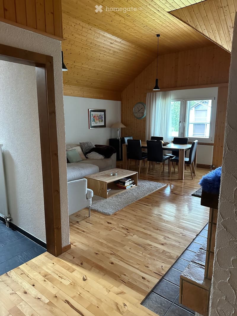 Bright, open living and dining area with wooden floors, a slanted wooden ceiling, and large windows. Features a sofa, coffee table, dining table with chairs, and a lamp.