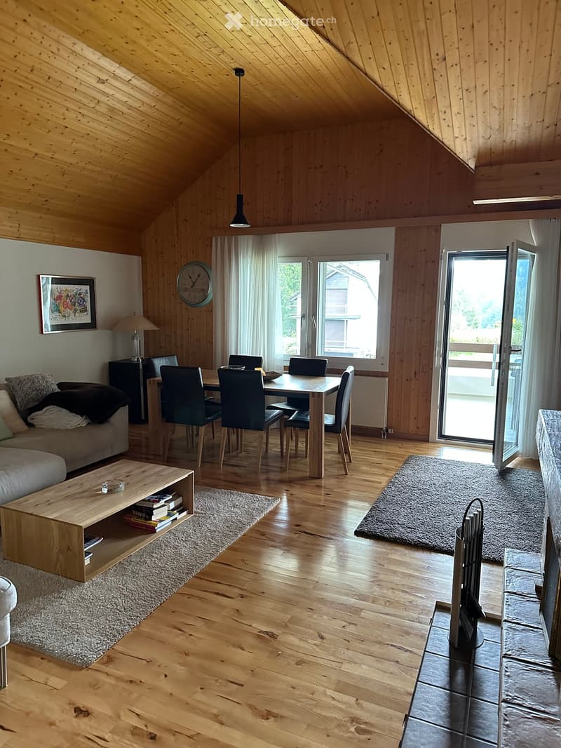 Bright, wooden-floored living and dining area with a sofa, coffee table, dining table, chairs, and a lamp. Large windows and a sliding glass door lead to a balcony.