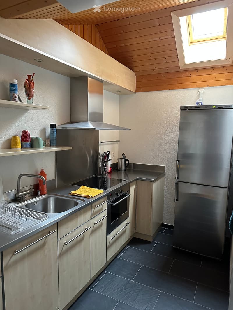 Modern kitchen with stainless steel appliances, wooden cabinets, and a dark tiled floor. Features include a sink, oven, refrigerator, and various kitchen utensils on the counter.
