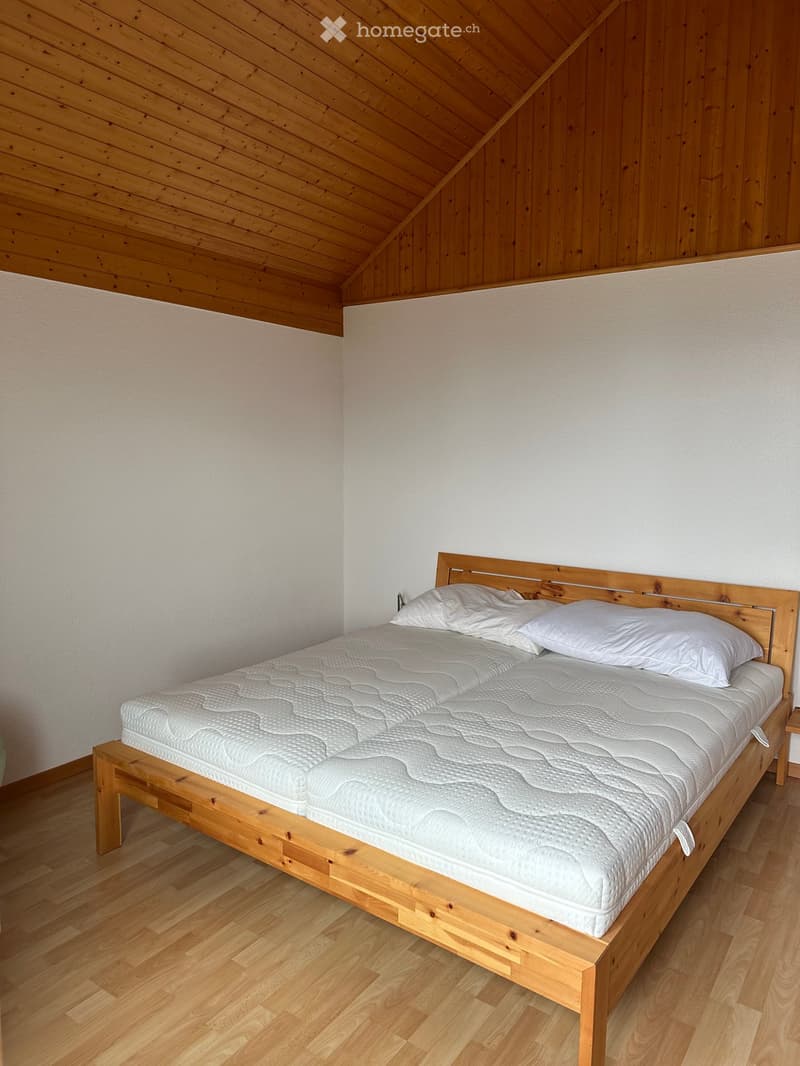A cozy bedroom with a wooden bed frame, a white mattress, and two white pillows. The room features light wooden walls and a slanted wooden ceiling.