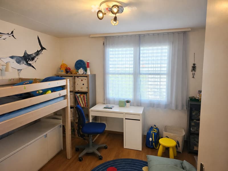 A well-lit bedroom with a bunk bed, desk, chair, bookshelf, and various toys and items. The room features a large window with blinds and a decorative shark mural on the wall.