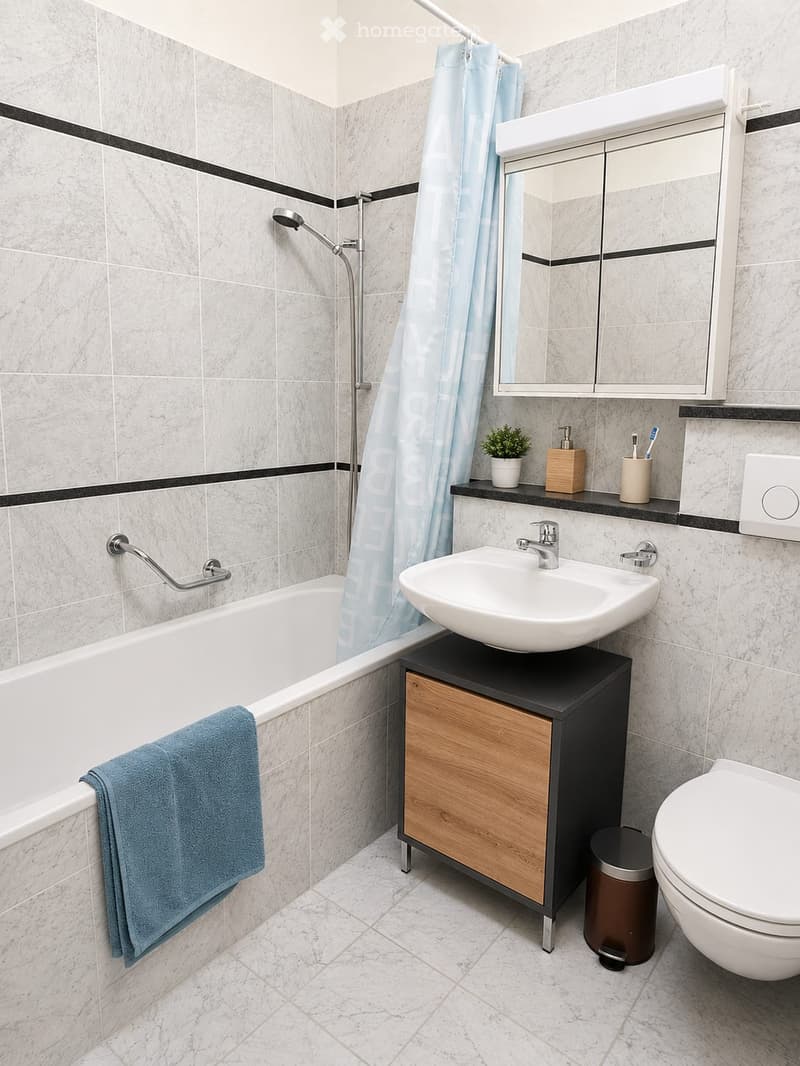 Modern bathroom with a bathtub, sink, toilet, and a mirrored cabinet. Features include a handheld shower, a towel on the edge of the tub, a small plant on the sink counter, and a trash can near the toilet.