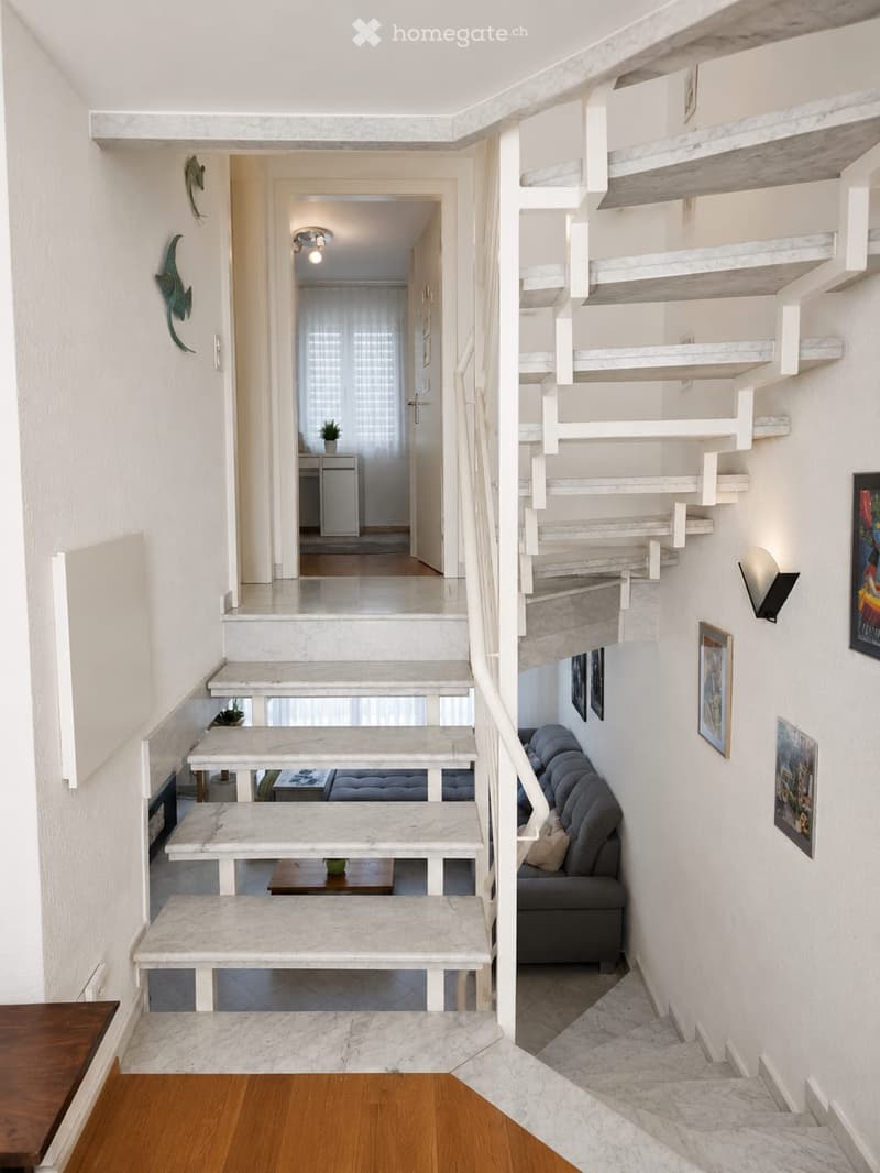 Modern hallway with white marble staircase, built-in shelves, and a small seating area with a sofa and coffee table. The space features contemporary decor with framed artwork and a small plant.