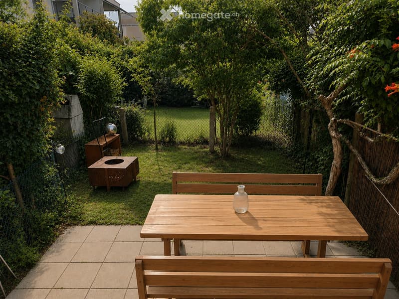 A small, well-maintained garden with a wooden table, two benches, and a decorative vase. The area is surrounded by lush greenery and features a stone pathway.