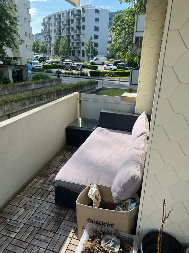 Balcony with wooden decking, a sofa with cushions, a cardboard box, and a plastic container with debris.