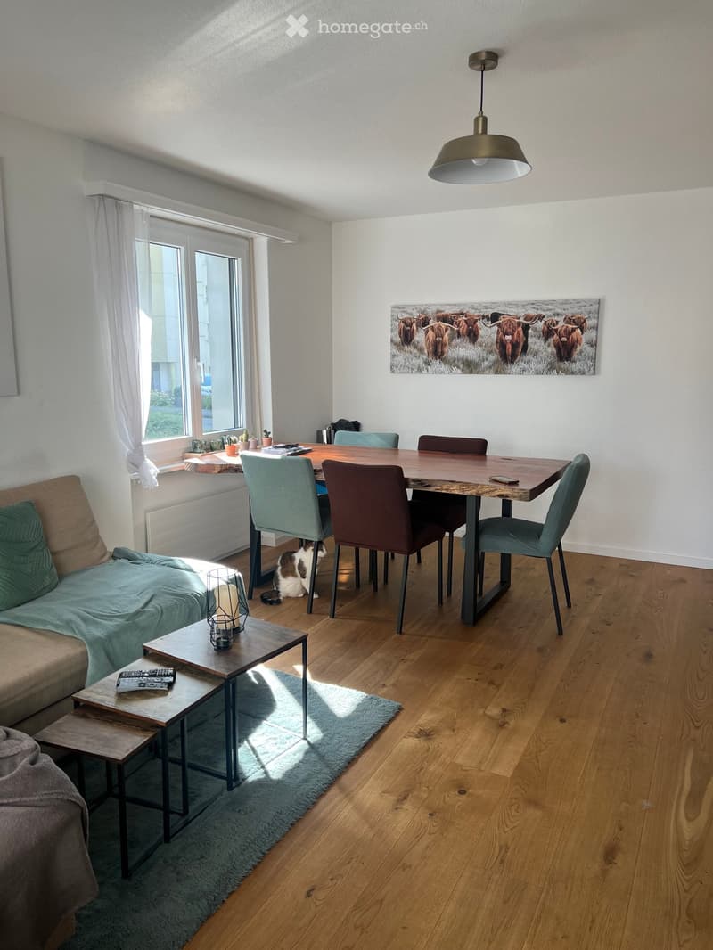 Living and dining area with a sofa, coffee table, dining table, chairs, and a cat. Features wooden flooring, large windows, and a painting on the wall.