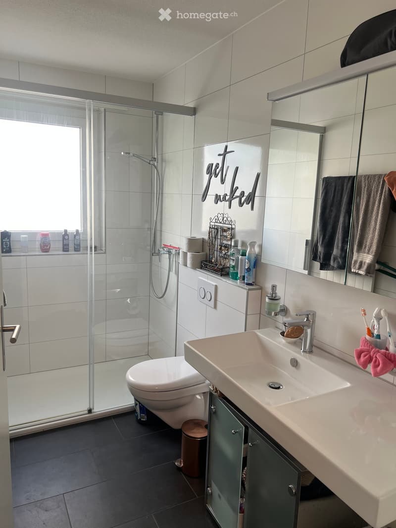 Modern bathroom with a white sink, mirror, toilet, and glass-enclosed shower. Includes towels, toiletries, and a decorative sign on the wall.