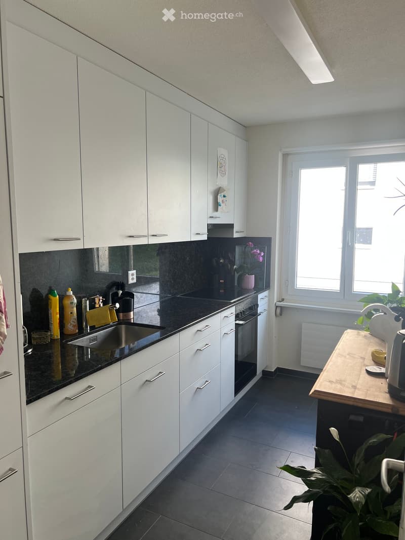 Modern kitchen with white cabinets, black countertops, stainless steel sink, and various appliances. Features a window with a view, a small plant, and a wooden cutting board with a kettle on it.