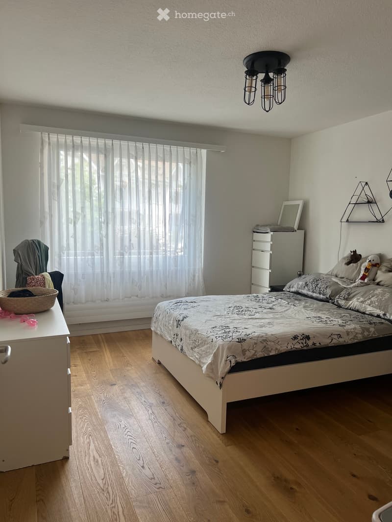 Cozy bedroom with wooden flooring, a double bed with patterned bedding, a nightstand with a mirror, and a wicker basket on a small table near the window.