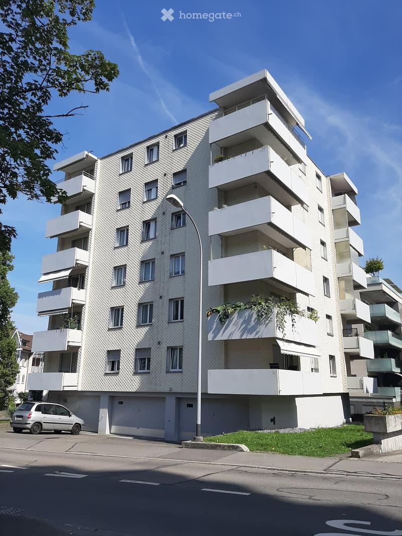 Modern multi-story apartment building with white balconies and textured facade, featuring multiple windows and a garage area at the base.