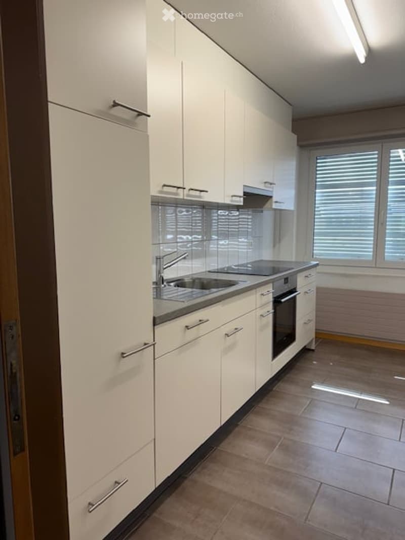 Modern kitchen with white cabinets, stainless steel sink, and oven. Features tiled flooring and large windows with blinds.
