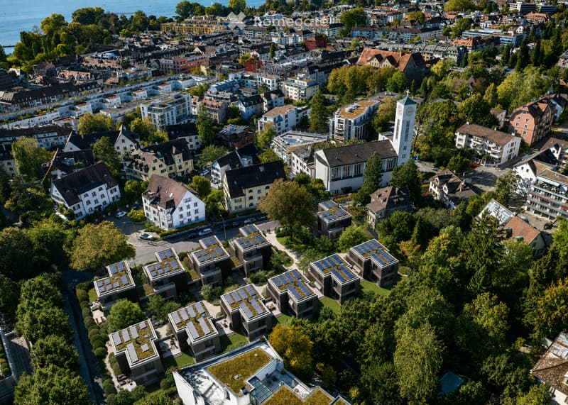 Aerial view of a dense urban area with numerous buildings, including residential and commercial structures, surrounded by greenery and trees. The area features a mix of architectural styles, with some buildings equipped with solar panels on their rooftops