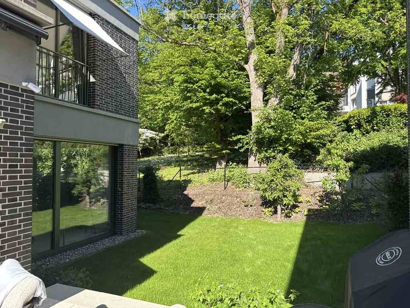 Well-maintained garden with a neatly trimmed lawn, surrounded by trees and shrubs. A modern house with a balcony is visible in the background.