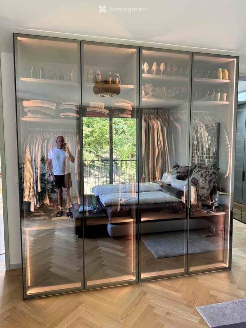 Modern bedroom with large mirrored wardrobe, neatly arranged clothing, and a stylish bed with decorative pillows.