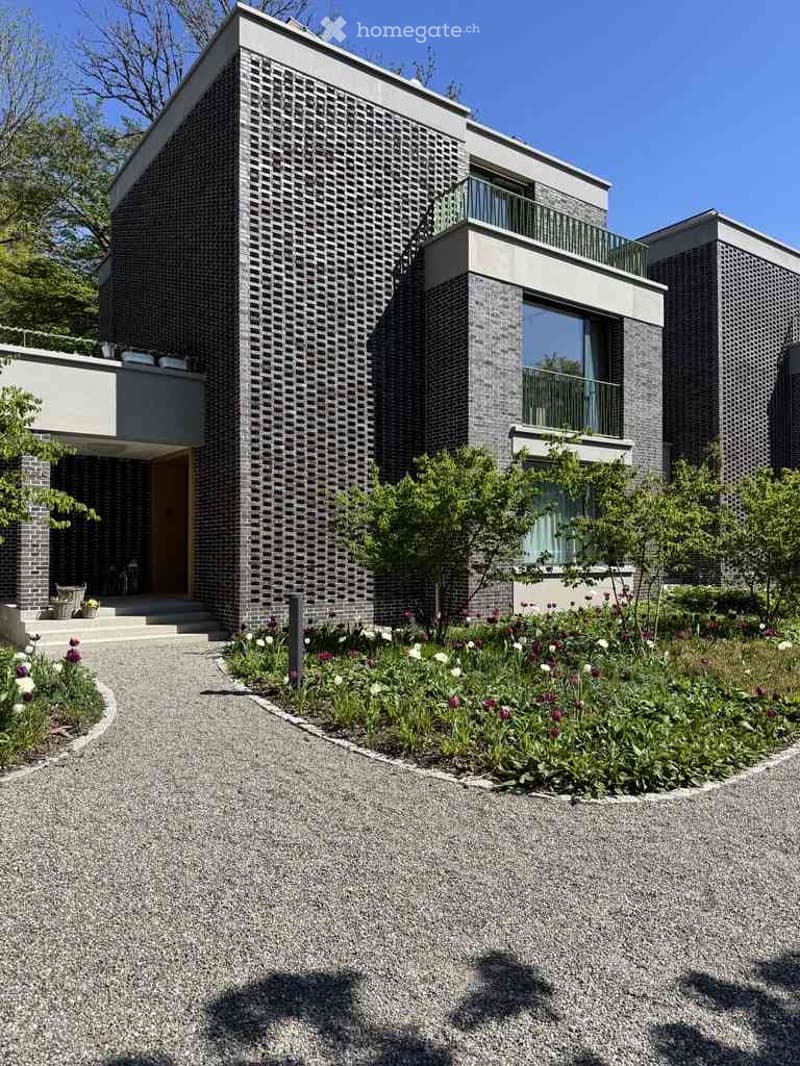 Modern two-story house with grey brick facade, large windows, and balconies. The front features a landscaped garden with flowers and a gravel driveway.