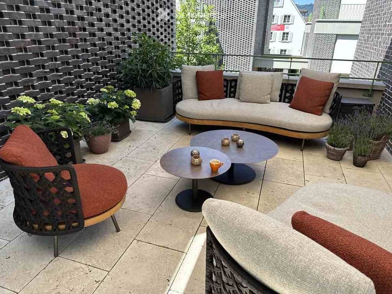 Modern balcony with a sofa, chairs, coffee tables, and potted plants, overlooking a cityscape.