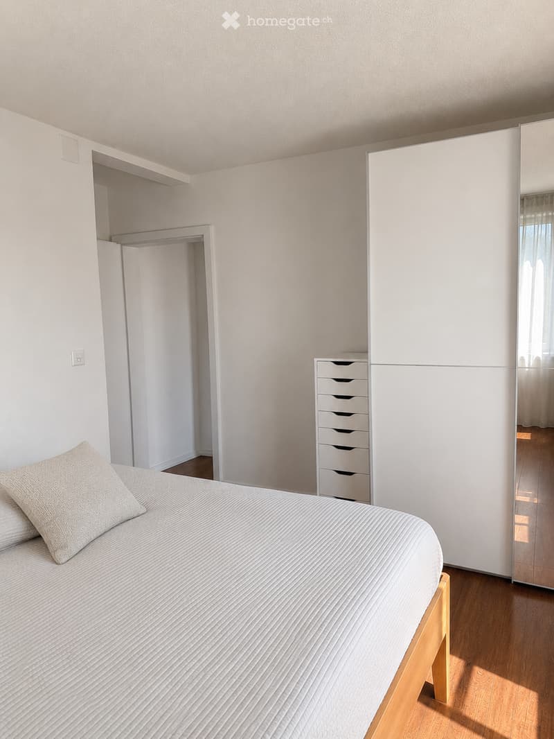 Minimalist bedroom with a white bed, light-colored pillows, a large white wardrobe with a mirror, and a small drawer unit.