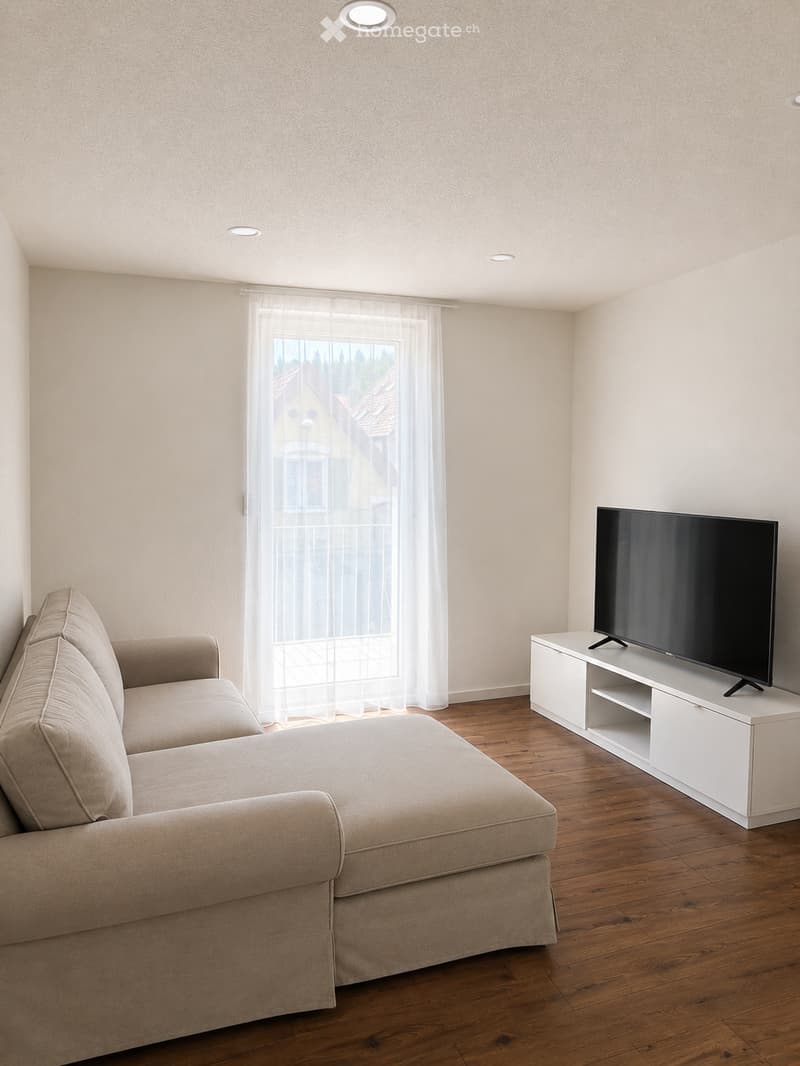 Modern living room with a beige sectional sofa, a flat-screen TV on a white stand, and large windows with sheer curtains.
