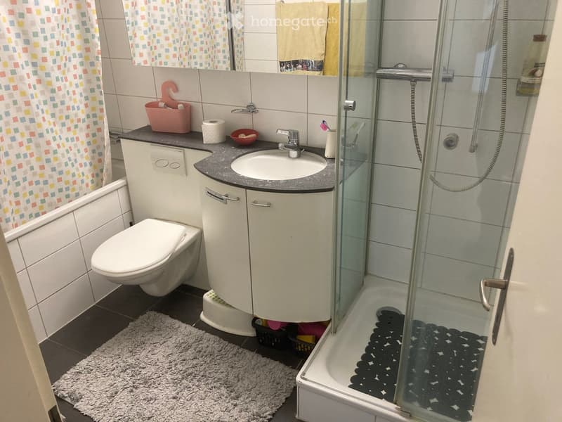 Small bathroom with shower, sink, and toilet. White tiled walls, gray floor, and mirror.
