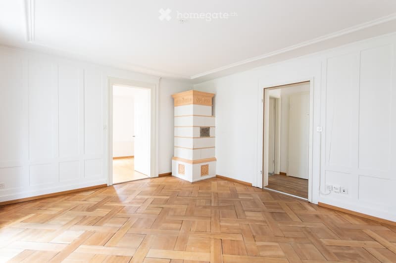 Empty room with white walls, wooden herringbone flooring, and two open doorways leading to other spaces. A decorative column is present in the center.