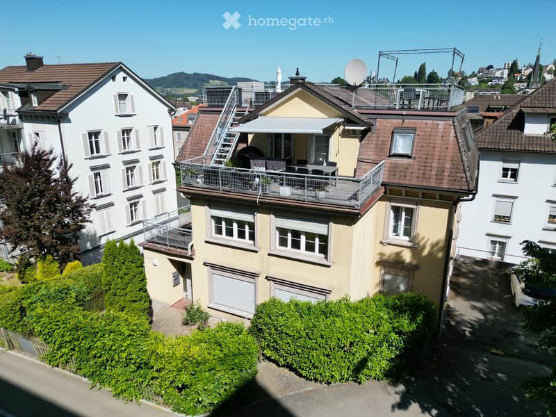 Multi-story residential building with beige and white exterior, multiple windows, balconies, and a rooftop terrace with outdoor furniture.