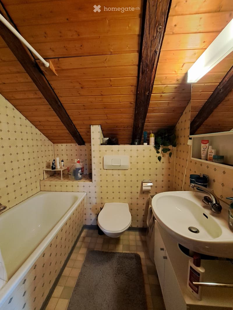 Cozy attic bathroom with wooden walls and ceiling, featuring a bathtub, toilet, sink, and small shelf with toiletries.