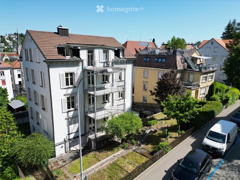 Multi-story residential building with balconies, surrounded by greenery and parked cars.