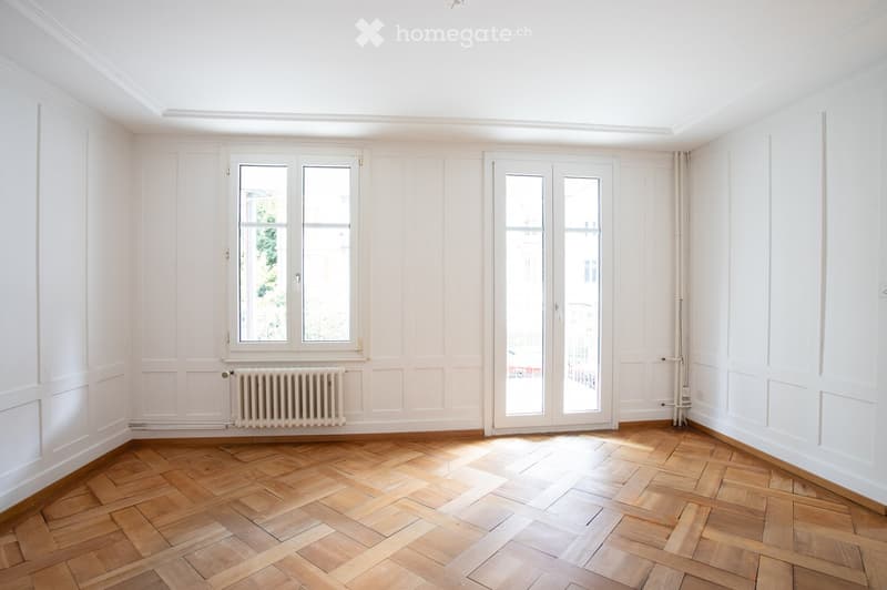 Empty room with white walls, wooden herringbone floor, large windows, and a radiator.