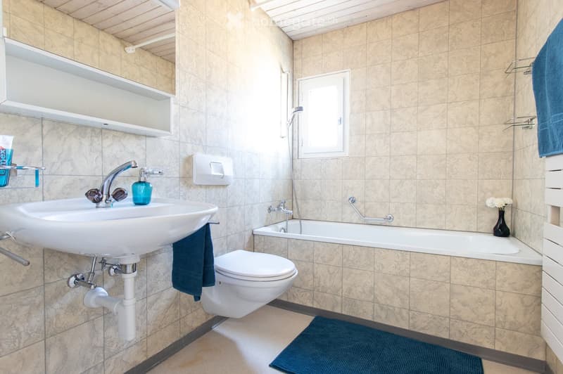 Modern bathroom with beige tiled walls and floor, featuring a white sink with a chrome faucet, a toilet, a bathtub with a showerhead, a mirror, and a blue towel on the floor.