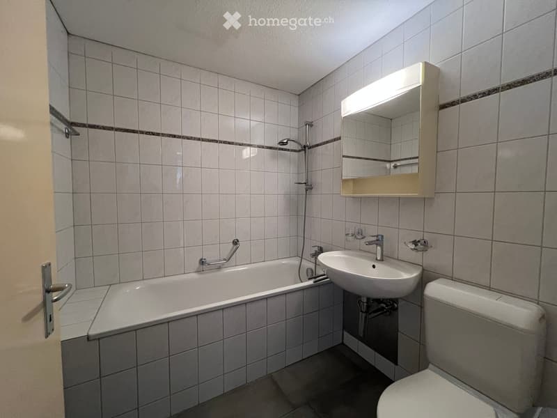 Bathroom with a bathtub, sink, toilet, mirror, and showerhead. The walls are tiled with a decorative border, and the floor is tiled.