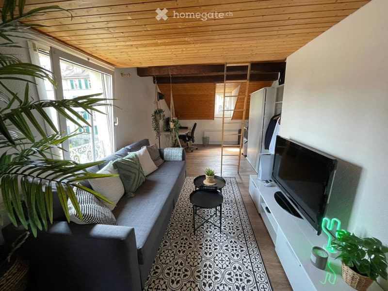 A modern, open-concept living space with a grey sectional sofa, patterned rug, coffee table, TV stand, and a small workspace area with a desk and chair. The room features wooden flooring and a wooden ceiling with exposed beams.