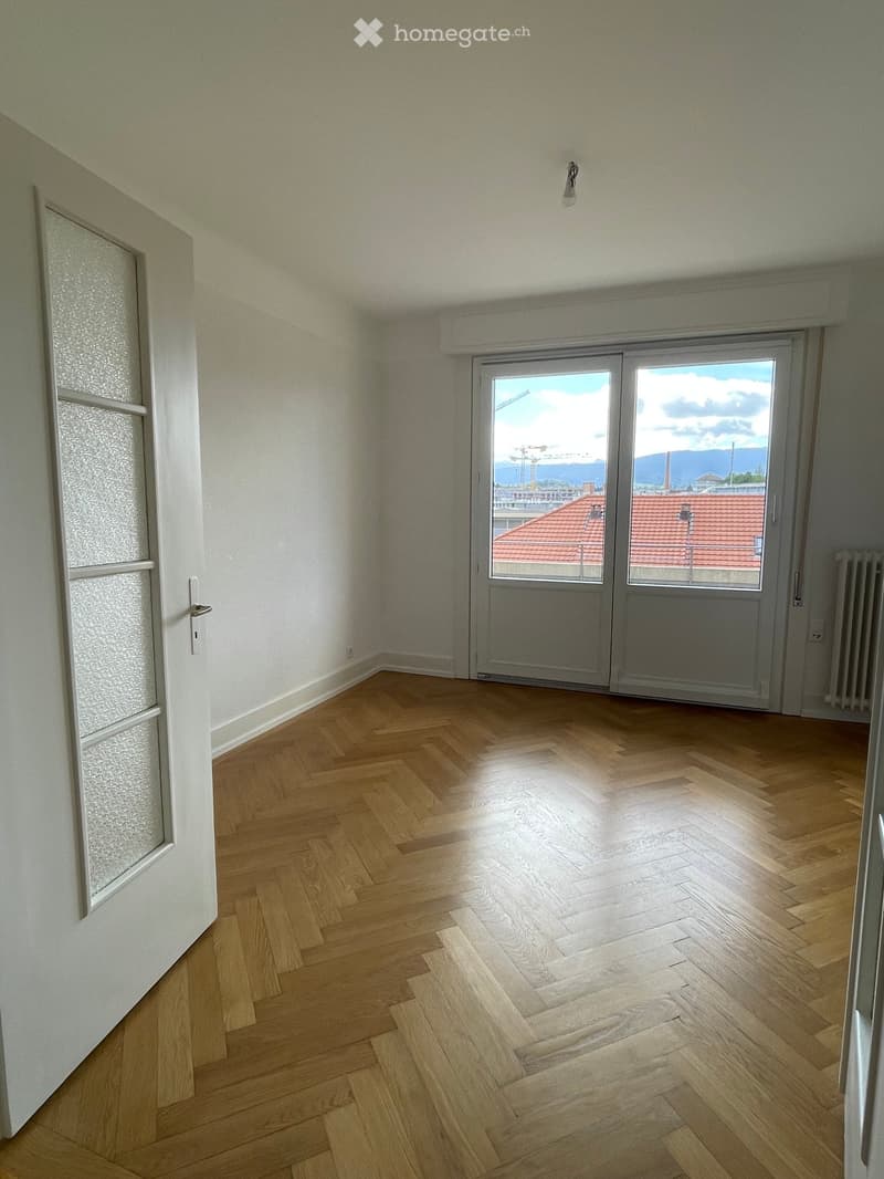 Empty room with wooden herringbone flooring, white walls, and large sliding glass doors leading to a balcony with a view of rooftops and mountains.