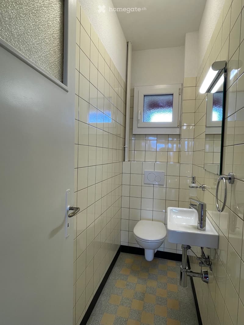 Modern bathroom with white tiled walls, a toilet, a sink with a chrome faucet, a mirror, and a small window with frosted glass. The floor features a checkered pattern in beige and grey.