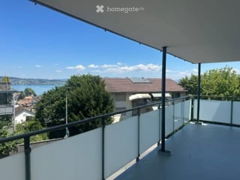 Balcony with glass railing, overlooking a lake and surrounding buildings.
