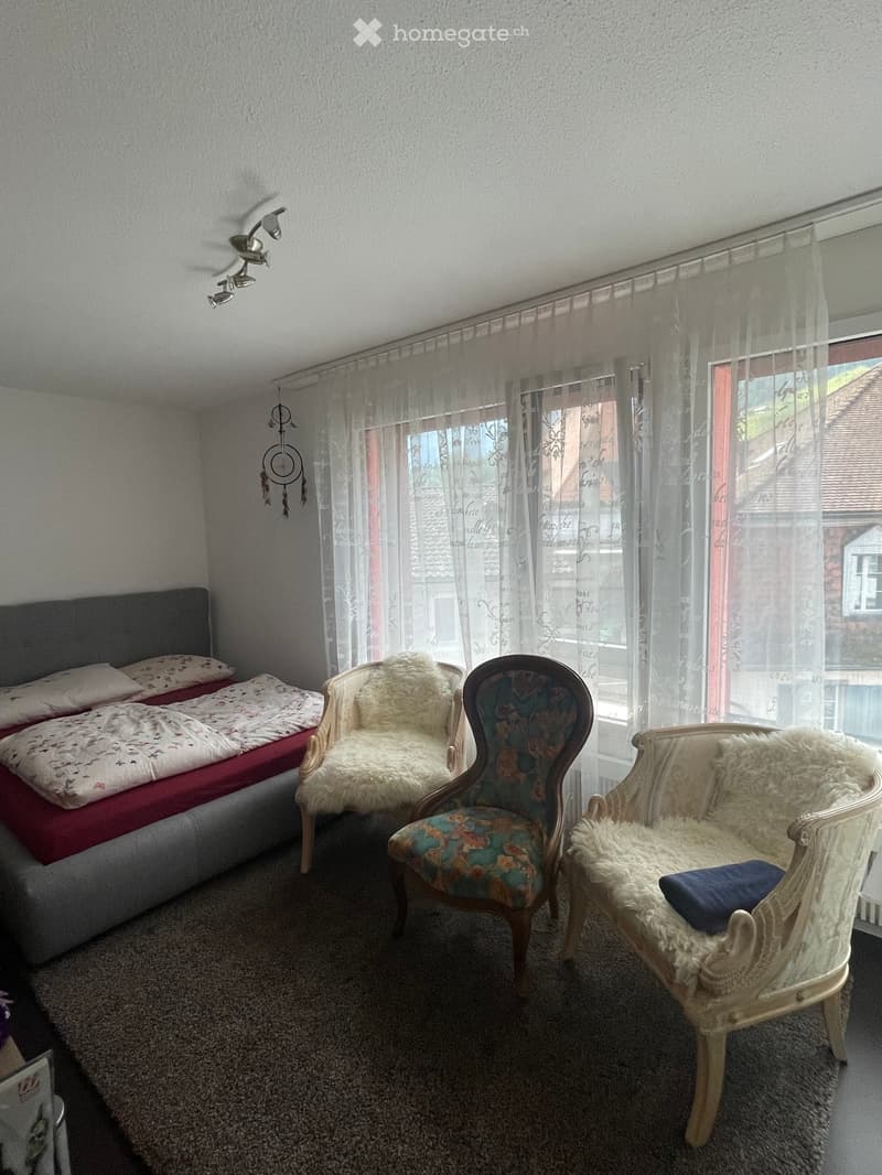 Cozy bedroom with a double bed, three chairs, and sheer curtains covering a large window. The room features a carpeted floor and decorative lighting.