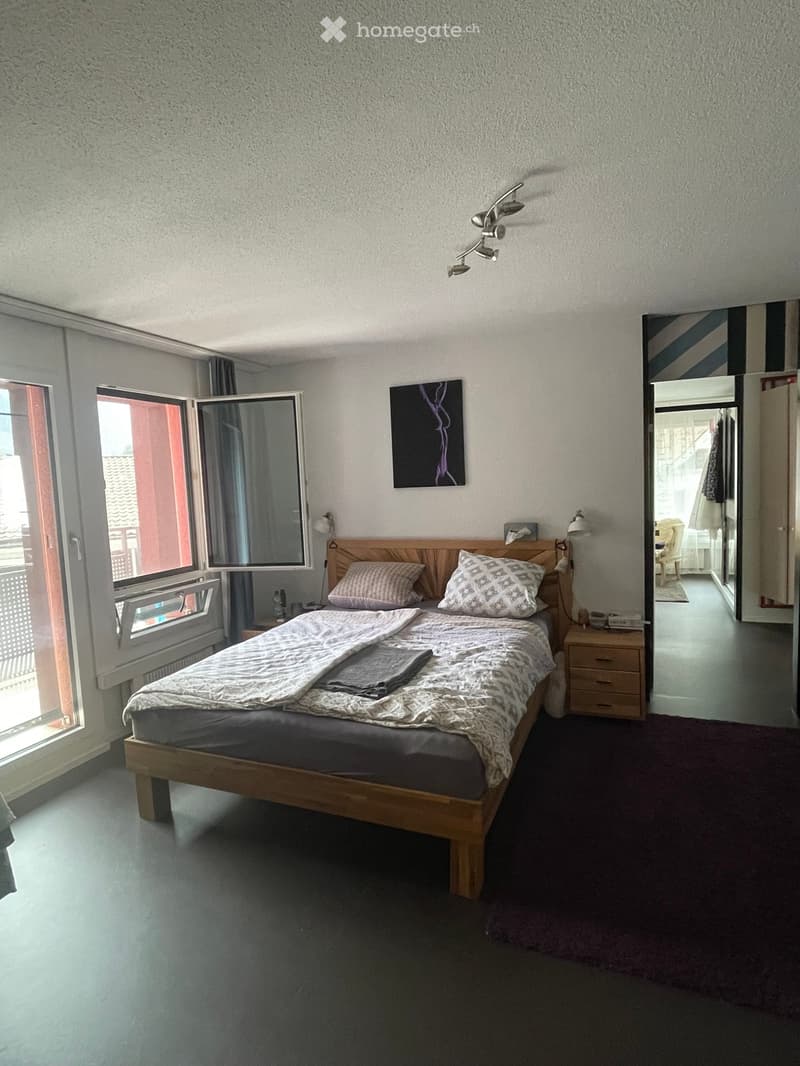 Cozy bedroom with a wooden bed, nightstand, mirror, and artwork on the wall. The room features large windows with open blinds and a view of the outside.