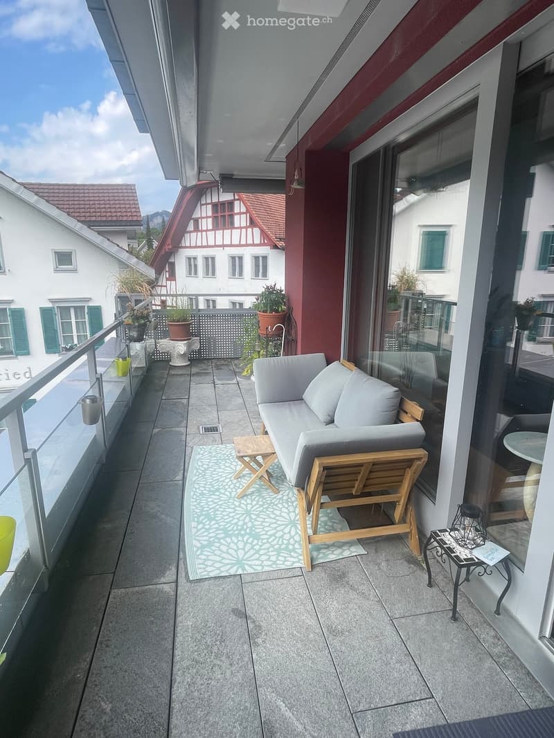 Balcony with a sofa, chair, small table, and rug, overlooking a town with traditional houses.