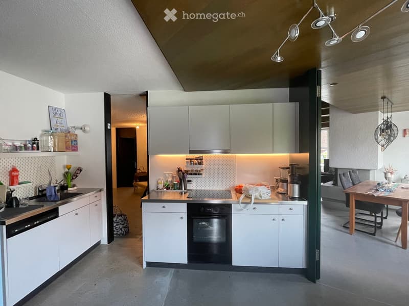 Modern kitchen with white cabinets, stainless steel appliances, and a central island. Visible items include a sink, oven, blender, and a small dining area with a table and chairs.