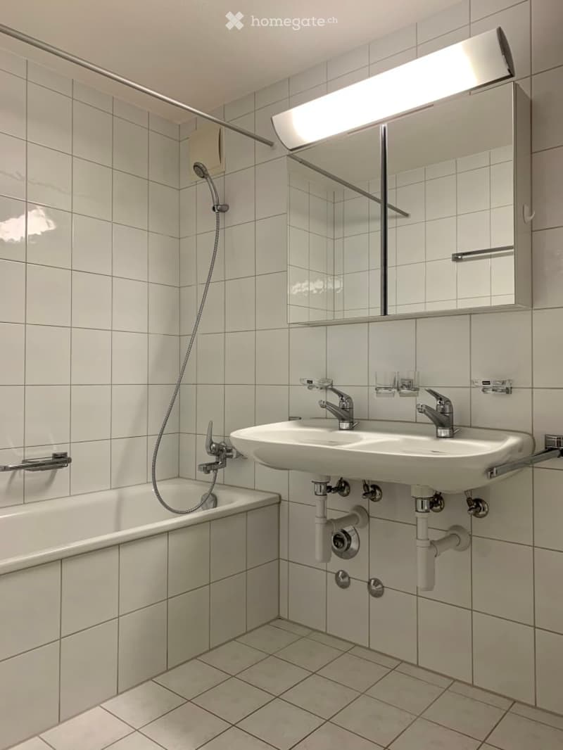 Modern bathroom with white tiled walls and floor, featuring a bathtub, a sink with dual faucets, a mirror with built-in lighting, and a handheld showerhead.