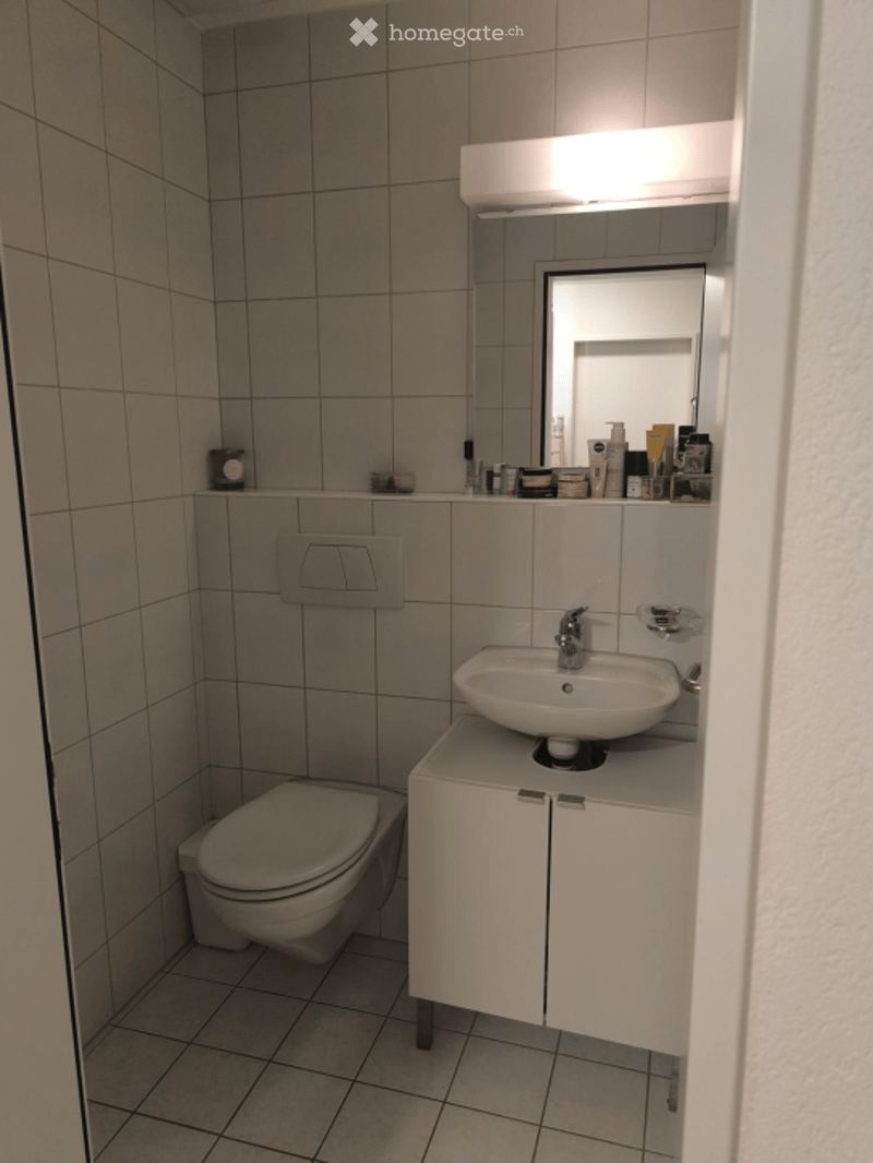 Modern bathroom with white tiles, a toilet, a sink with a cabinet, and a mirror above the sink. The cabinet has a sleek design with a silver handle.