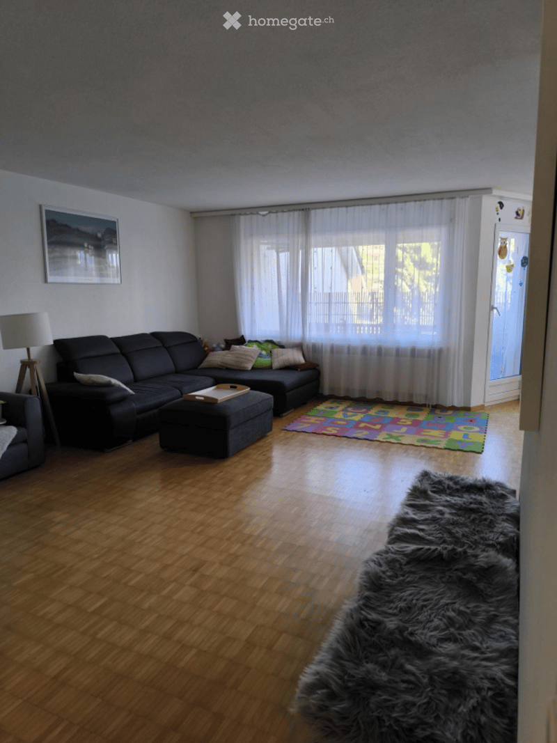 Living room with hardwood flooring, large window with white curtains, black sectional sofa, ottoman, floor lamp, picture frame, colorful play mat, and a furry rug.