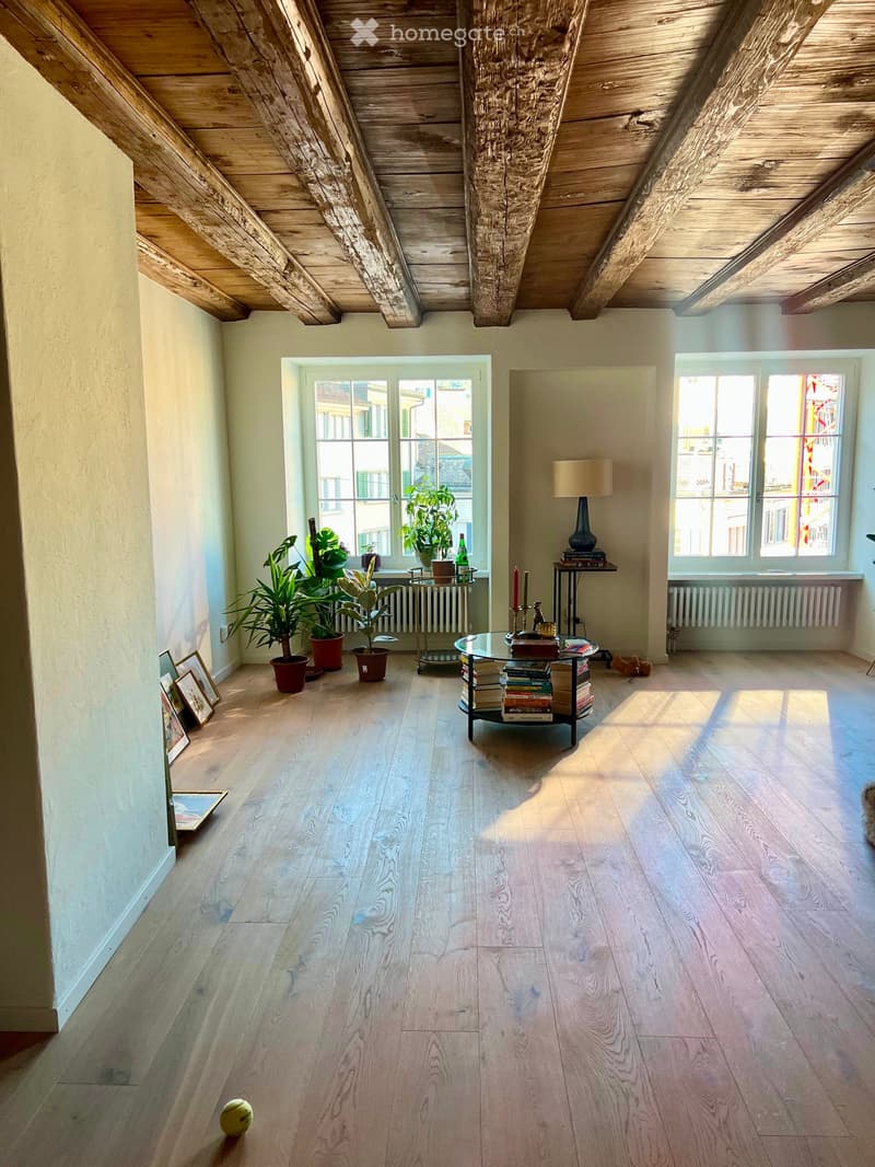 Bright, airy room with wooden floors and exposed wooden beams on the ceiling. Features large windows, a glass coffee table, potted plants, a lamp, and picture frames on the wall.
