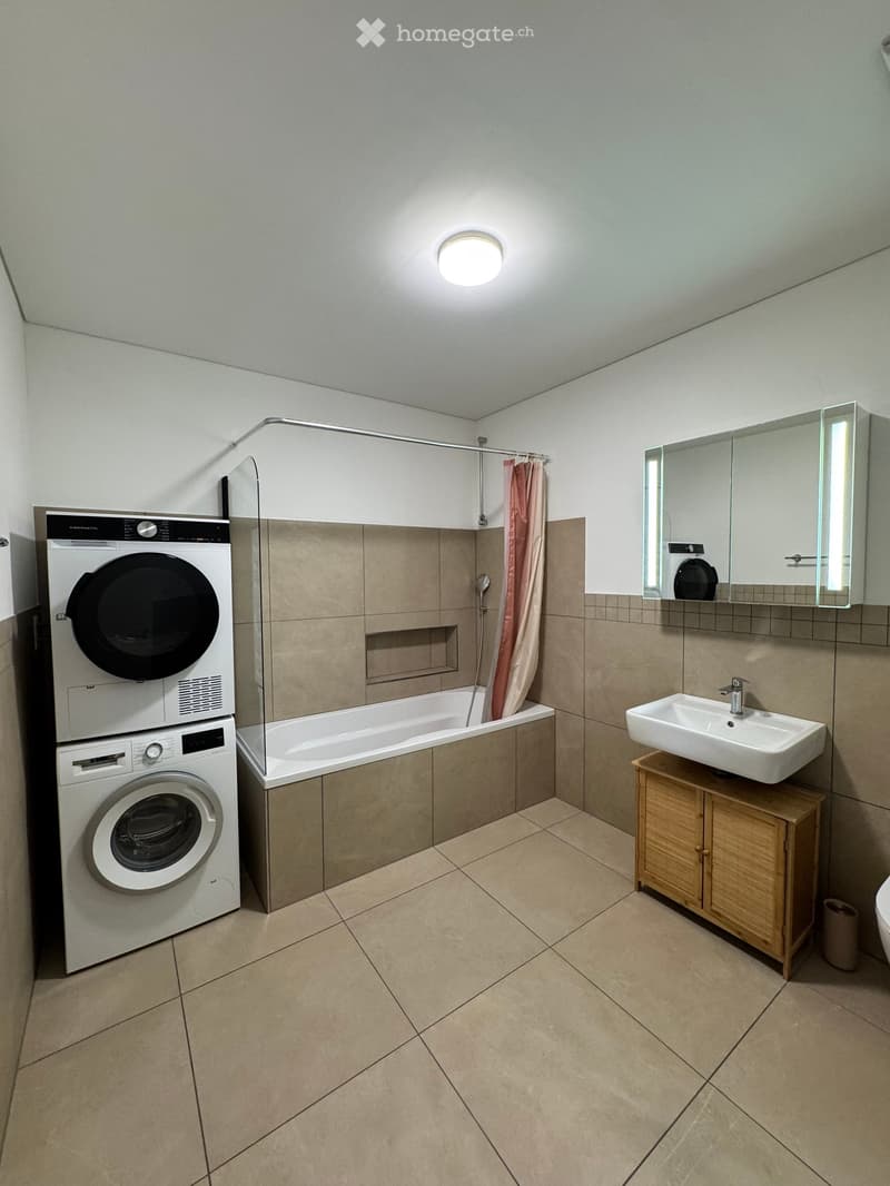Modern bathroom with beige tiles, a bathtub, a sink with a wooden cabinet, a mirror, and a stacked washing machine and dryer.