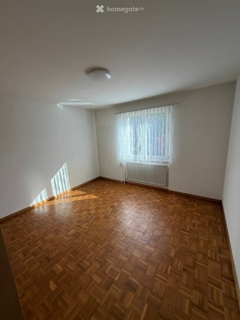 Empty room with wooden parquet flooring, white walls, a single window with white curtains, and a radiator below the window.