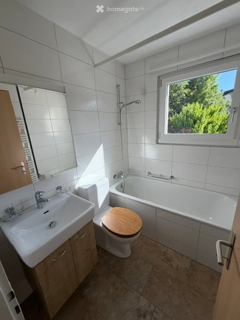Modern bathroom with white tiles, a wooden toilet seat, a bathtub, a sink with a mirror, and a window offering a view of greenery.