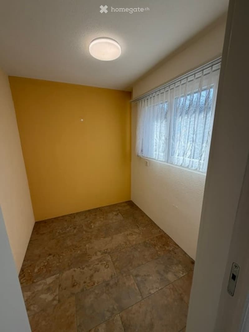 Empty room with beige walls, a single window with white blinds, and a tiled floor. The room has a round ceiling light fixture and a door visible on the right side.