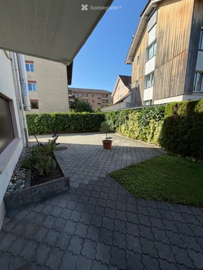 A paved outdoor area with a small garden bed, a potted plant, and surrounding buildings.