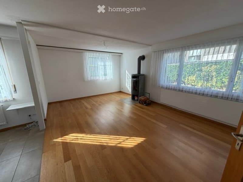 Empty room with wooden flooring, white walls, large windows with curtains, and a fireplace in the corner.