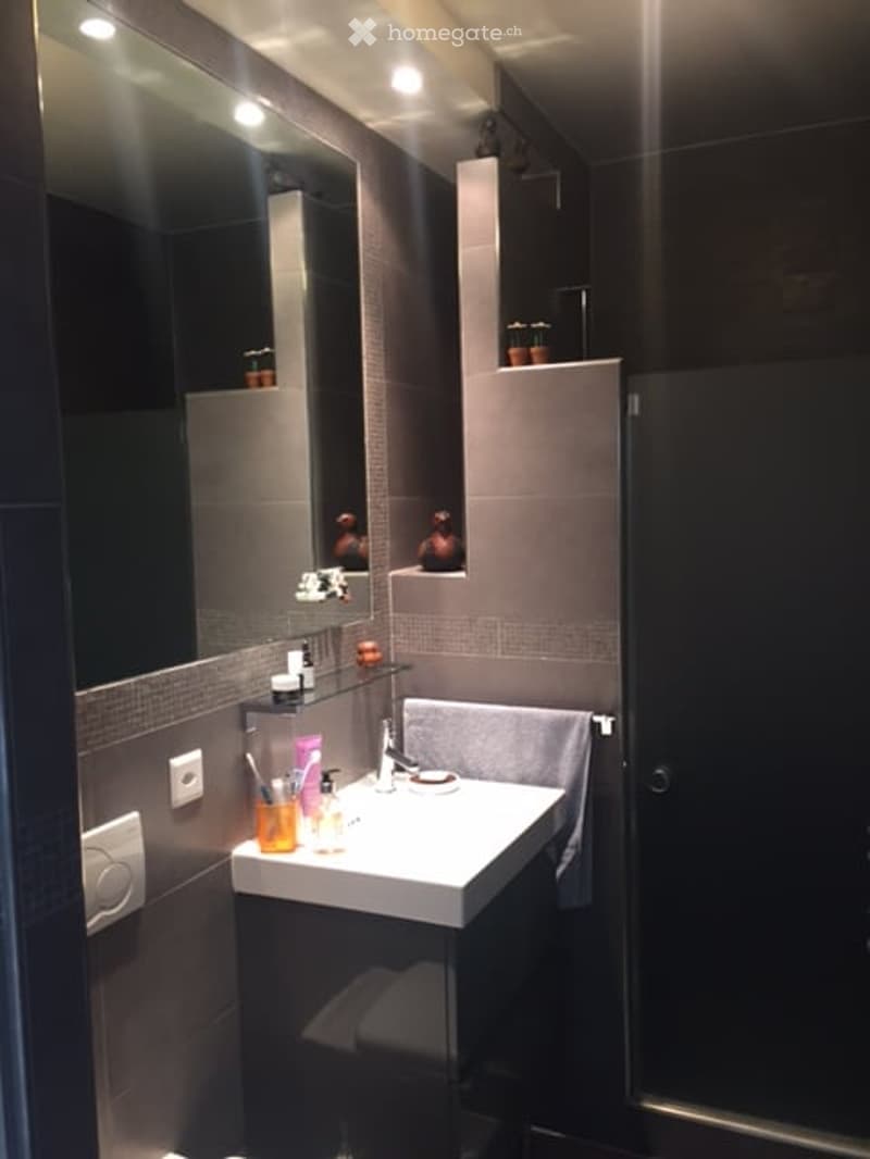 Modern bathroom with a white sink, large mirror, and grey tiled walls. The sink has a chrome faucet and is surrounded by various toiletries and a folded towel on a rack.